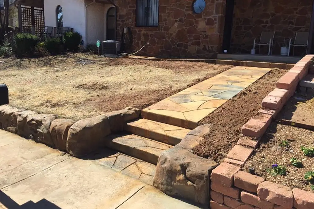 A rustic front yard with a stone walkway and steps leading to a house, bordered by a natural stone retaining wall and a brick-edged garden bed.