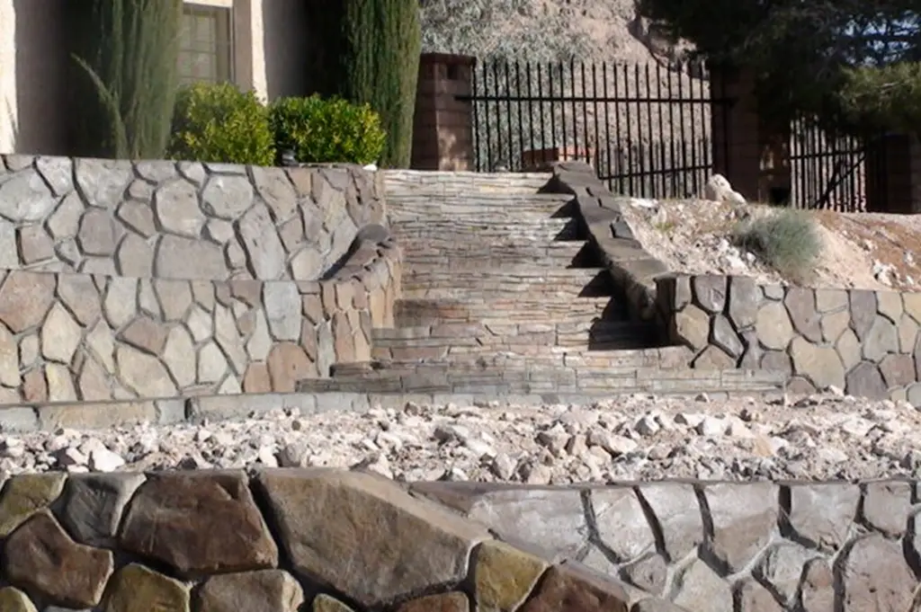A set of stone steps leading to a gated entrance, surrounded by stone retaining walls with a mix of irregular and uniform patterns. The hardscape design integrates natural stone elements with structured landscaping.